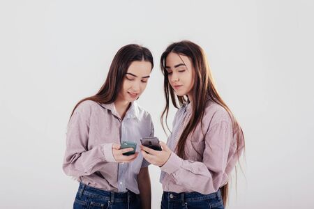 Sharing some information using smartphones. Two sisters twins standing and posing in the studio with white background.の写真素材