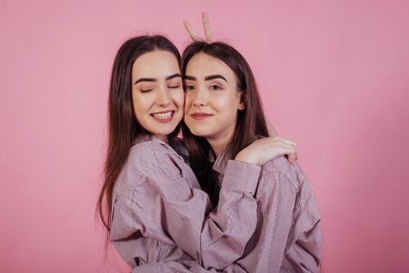 Girl from the left makes horn gesture behind other woman. Two sisters twins standing and posing in the studio with white background.の写真素材