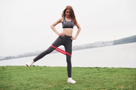 Exercise for legs. Fitness girl have morning workout at cloudy day near the lake with special red...