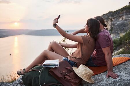 Hugging couple with backpack sitting on top of rock mountain enjoying view coast a river or lake. Traveling mountains and coast, freedom and active lifestyle concept.の写真素材