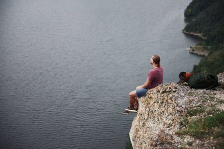 Traveller with backpack sitting on top of mountain enjoying view above the water surface. Traveling along mountains and coast, freedom and active lifestyle concept.の写真素材