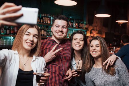 Guy shows gesture of two fingers. Friends taking selfie in beautiful nightclub. With drinks in the hands.の写真素材