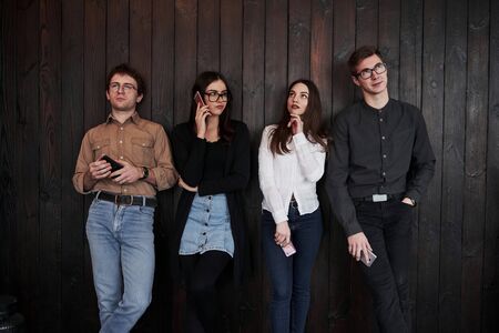 Everyone looks in different directions. Youth stands against black wooden wall. Group of friends spending time together.の写真素材