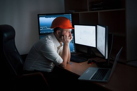 In the safety helmet. Bearded man in white shirt and red safety helmet works in the office with multiple computer screens in index charts.の写真素材