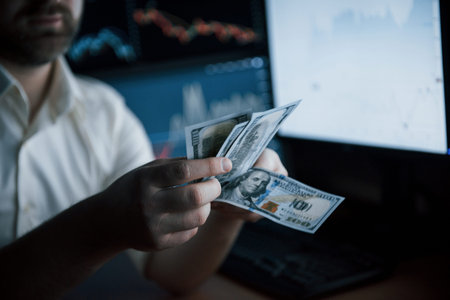 Its payday. Bearded man in white shirt counts money in the office with multiple computer screens in index charts.の写真素材
