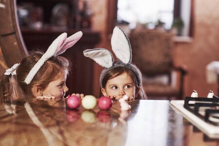 Cute kids. Funny two sisters in bunny ears hidding near the table with painted eggs.の写真素材