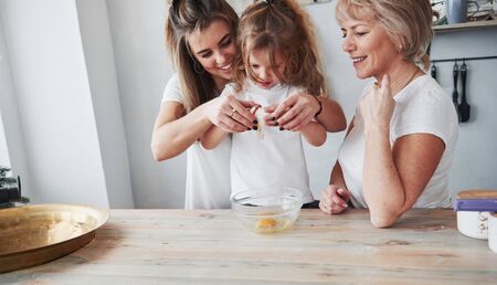 Picture in motion of breaking egg. Mother, grandmother and daughter having good time in the kitchen.の写真素材