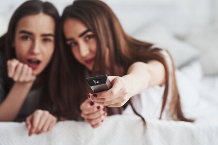 Black remote controller accented in photo. Two female twins lying on the white bed at daytime watching movie.の写真素材