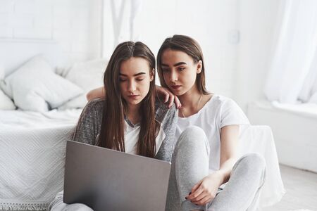 Seriously looking girls. Young female twins sitting on the floor near white bed and using silver colored laptop.の写真素材
