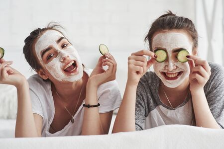 Nice day. Conception of skin care by using fresh cucumber rings and white mask on the face. Two female sisters have weekend at bedroom.の写真素材