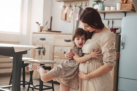 Pregnant woman standing and hugs her daughter at the kitchen at daytime.の写真素材
