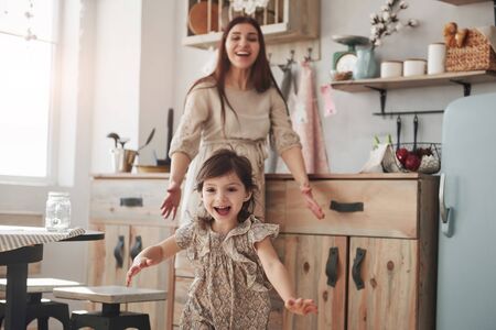 Be happy like this little girl. Playful female child have fun by running in the kitchen at daytime of front of her mother.の写真素材