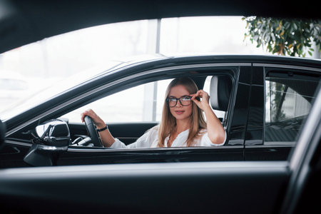 Against big windows. Beautiful blonde girl sitting in the new car with modern black interior.