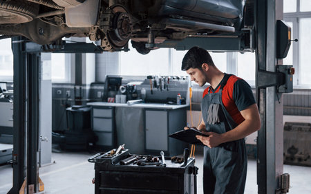 Nice looking autosalon. Man at the workshop in uniform using notepad for his job for fixing broken...