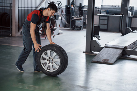In red and black colored uniform. Mechanic holding a tire at the repair garage. Replacement of...