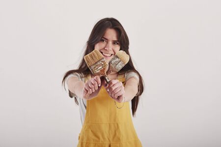 Holding brushes in hands. Brunette woman in yellow uniform stands against white background in the studio.の写真素材