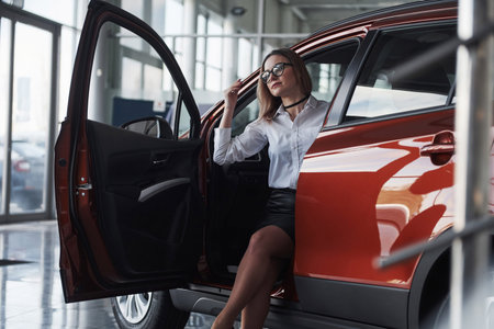 Opening the door. Woman in eyewear, black skirt and white shirt sits in the red automobile.