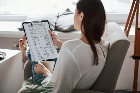 Looking at the plan of a building. Young brunette in the room with white walls and daylight that...