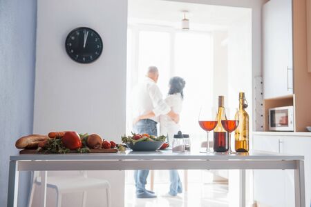 Waiting for the right time and looking through the window. Senior man and his wife in white shirt have romantic dinner on the kitchen.の写真素材