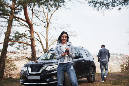 Holds cup with warm drink. Couple have arrived to the forest on their brand new black car.