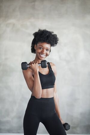 Having fun during process. Studio shot of young woman that stands in front of grey background with dumbbells in hands.の写真素材