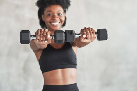 Showing equipment. Studio shot of young woman that stands in front of grey background with dumbbells in hands.の写真素材