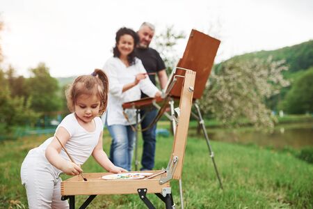 Wooden easel. Grandmother and grandfather have fun outdoors with granddaughter. Painting conception.の写真素材