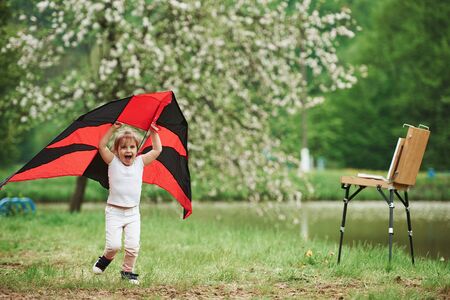 Scream of happiness. Positive female child running with red and black colored kite in hands outdoors.の写真素材