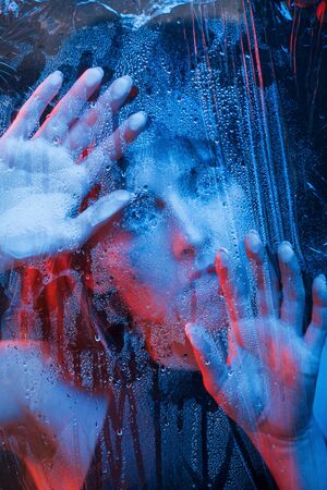 Cool temperature. Studio shot in dark studio with neon light. Portrait of beautiful girl behind wet glass.の写真素材