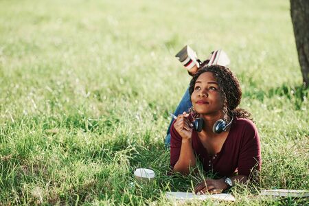 Writer thinks how to finish the chapter. Cheerful african american woman in the park at summertime.の写真素材
