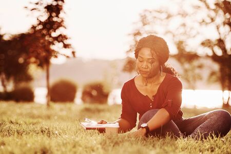 Takes needed items for writing. Cheerful african american woman in the park at summertime.の写真素材