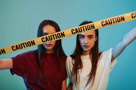 Behind yellow caution tape. Studio shot indoors with neon light. Photo of two beautiful twins.の写真素材