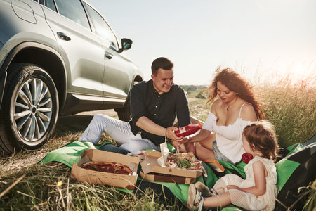 Family have picnic at countryside near silver automobile at sunset.