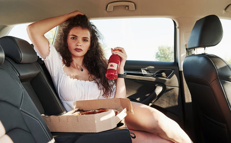 Brunette with curly hair sits in the modern car with bottle with tomato juice in hand and pizzas.