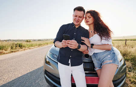 Using smartphone. Lovely couple near their new modern car at weekend time.の写真素材