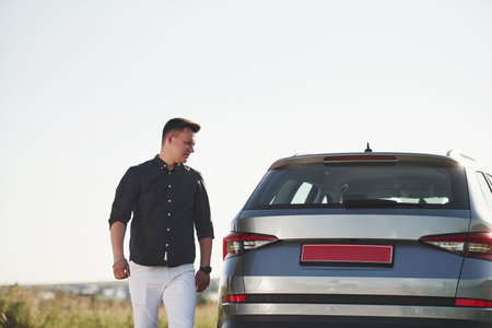 Man in elegant clothes walks near his silver colored car at sunny day.
