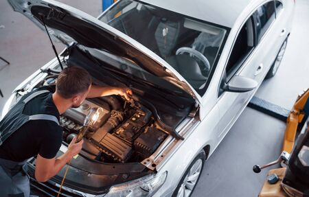 With flashlight. Man in grey uniform repairs white automobile indoors.の写真素材