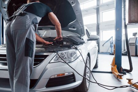 Checks voltage. Man in grey uniform repairs white automobile indoors.の写真素材