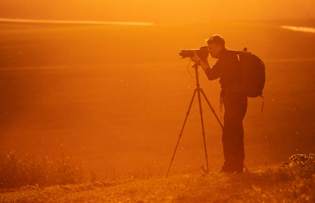 Photographer with professional equipments makes photos. Stands in the field illuminated by sunlight.
