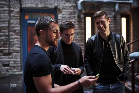 With smartphone. Some funny stuff. Three male friends stands outside the bar. Having conversation. In nice clothes.の写真素材