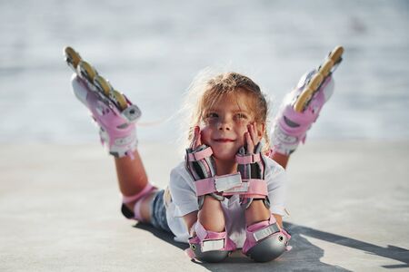 Lying down. Cute little girl with roller skates outdoors near the lake at background.の写真素材