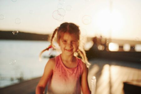 Amazing sunlight. Happy little girl playing with bubbles near the lake at park.の写真素材