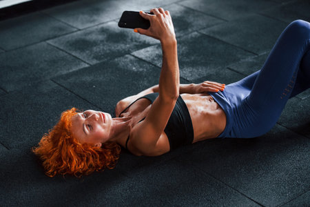Taking a selfie with phone. Sporty redhead girl have fitness day in gym at daytime. Muscular body...