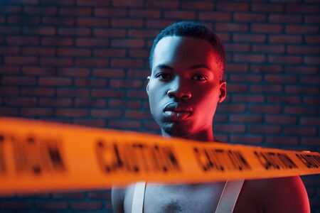Yellow caution tape and uniform. Futuristic neon lighting. Young african american man in the studio.の写真素材