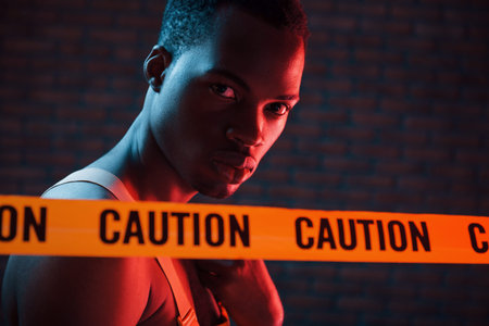 Yellow caution tape and uniform. Futuristic neon lighting. Young african american man in the studio.