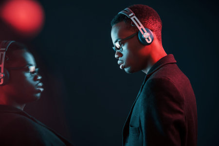 Enjoying listening music in headphones. In glasses. Futuristic neon lighting. Young african american man in the studio.の写真素材