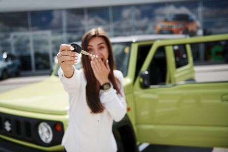 Cheerful emotions. Holds keys of her new vehicle. Young woman in white official clothes stands in front of green automobile outdoors.の写真素材