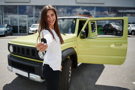 Holds keys of her new vehicle. Young woman in white official clothes stands in front of green automobile outdoors.の写真素材