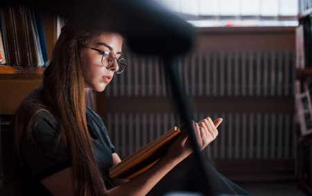Concentrated at reading. Female student is in library that full of books. Conception of education.