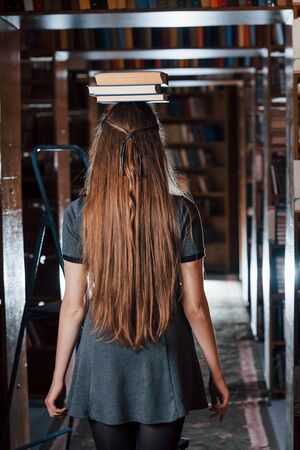 Cheerful girl have fun while puts books on her head. Female student is in library. Conception of education.の写真素材
